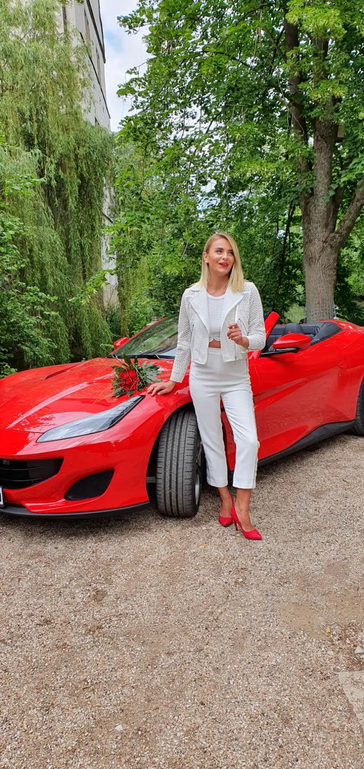 Braut im Zweiteiler aus Spitzentop, weißer Hose und Jacke vor einem roten Cabrio